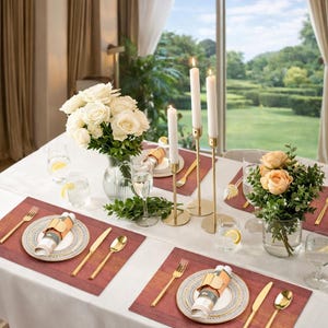 Puede incluir: Una mesa de comedor elegantemente decorada con rosas blancas y flores color melocotón en jarrones de cristal. Candelabros dorados con velas encendidas, cubertería dorada y platos estampados sobre manteles individuales rojos.