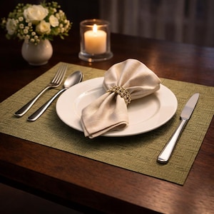 Puede incluir: Una mesa formal con un plato blanco, cubiertos y una servilleta beige doblada sujeta por un anillo dorado. Un mantel individual verde, un pequeño jarrón con flores blancas y una vela encendida en un vaso completan el arreglo.