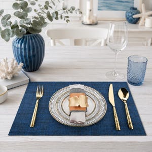 Puede incluir: Una mesa puesta con un mantel individual azul marino, cubiertos dorados y un plato decorativo. Un servilletero dorado sujeta una servilleta gris y naranja. También hay un vaso azul y una copa de vino.