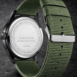 May include: Close-up of a black wristwatch with a green nylon strap. The watch face is silver with black hands and markers. The back of the watch is silver with the text "QUARTZ 5ATM STAINLESS STEEL" and "NO:0550".