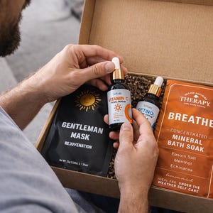 May include: A gift box containing skincare products. The box includes a black face mask labeled "Gentleman Mask", two serum bottles, and a packet of "Breathe" mineral bath soak. The Vitamin C and Retinol serum bottles have gold accents.