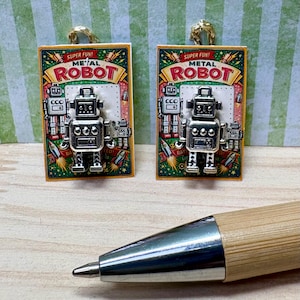 Op de afbeelding: Twee rechthoekige hangers met een retro robotontwerp. Elke hanger heeft een zilveren metalen robot op een kleurrijke achtergrond met de woorden "SUPER FUN! METAL ROBOT". De hangers hebben goudkleurige lussen aan de bovenkant.