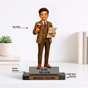 May include: A custom bobblehead figurine of a person in a brown suit, vest, and tie, holding a glass of dark beer and a newspaper. The figure stands on a black base with a grey plaque reading BLOKE THE LOCAL and a brown plaque reading MICHAEL COLLINS.