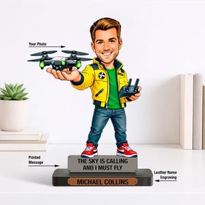 May include: A figurine of a man holding a drone and remote control. The man is wearing a yellow jacket, green shirt, blue jeans, and red sneakers. The base has the text "THE SKY IS CALLING AND I MUST FLY" and "MICHAEL COLLINS".