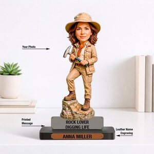 Pode incluir: Uma figura bobblehead personalizada de um geólogo com traje de safari bege, chapéu e botas, segurando um martelo de rocha. A figura está sobre uma base rochosa com uma placa cinza ROCK LOVER DIGGING LIFE e uma placa com ANNA MILLER.