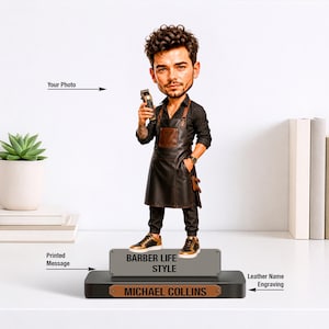 May include: A barber figurine holding clippers, dressed in a black leather apron and gold-detailed trainers. The base displays "BARBER LIFE STYLE" and "MICHAEL COLLINS". A succulent plant in a white pot and books are in the background.