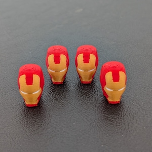 May include: Four red and gold Iron Man helmet figurines. The helmets are a vibrant red with gold faceplates, arranged on a dark surface. The design is a recognizable superhero icon.