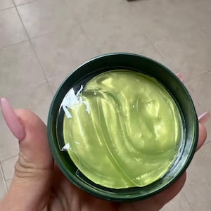 Op de afbeelding: Een ronde, donkergroene houder met meerdere doorschijnende, limoengroene gel-oogmaskers. De maskers zijn gestapeld in de ronde pot en hebben een gladde, vochtige en geleiachtige textuur die de gehele binnenkant van de houder opvult.