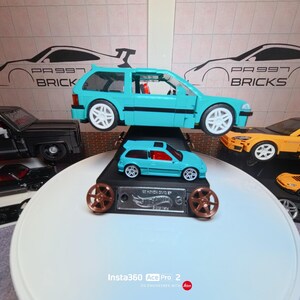 May include: A light blue brick-built hatchback car model displayed above a smaller light blue die-cast metal Honda Civic Si toy car. The display stand features a silver Hot Wheels logo plate and two bronze-colored wheel accents on the front.