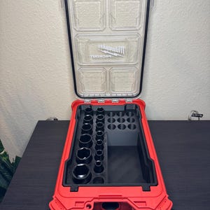 Puede incluir: Maleta de herramientas roja y negra abierta con tapa transparente. La maleta contiene una variedad de llaves de vaso negras y compartimentos de almacenamiento. La tapa tiene la palabra "Milwaukee PACKOUT" impresa.
