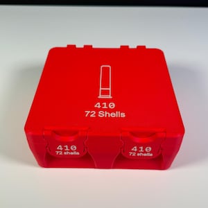 May include: A bright red plastic container for 410 shotgun shells. The container has a rectangular shape with a hinged lid. The text "410 72 Shells" is printed in white on the lid and sides, along with a graphic of a shell.