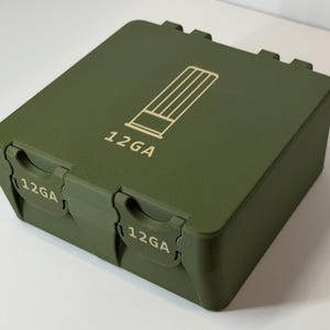 May include: An olive green ammunition box with the text "12GA" printed on the front and sides. The box has a rectangular shape with a hinged lid and two latches. A graphic of a shotgun shell is on the lid.
