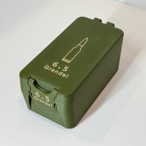 May include: An olive green ammunition box with a hinged lid. The box features a gold outline of a bullet and the text "6.5 Grendel" in gold. The box is rectangular with rounded edges and a textured surface.