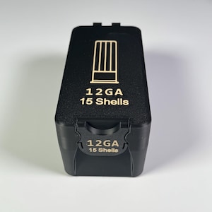 Op de afbeelding: Zwarte rechthoekige container met de tekst "12 GA 15 Shells" en een afbeelding van verticale lijnen. De container heeft een scharnierend deksel en is ontworpen om hagelpatronen in te bewaren. De container is gemaakt van matzwart materiaal.