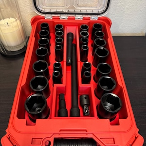 Könnte beinhalten: Eine rote Werkzeugkiste mit einem transparenten Deckel, gefüllt mit schwarzen Steckschlüsseln und Verlängerungen. Die Steckschlüssel sind in Reihen angeordnet, wobei verschiedene Größen sichtbar sind. Die Werkzeugkiste ist geöffnet und zeigt die geordnete Anordnung der Werkzeuge.