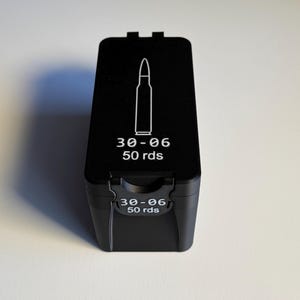 May include: A black ammunition box with white text and a bullet graphic. The text reads "30-06 50 rds" on the top and front of the box. The box is rectangular with a hinged lid.