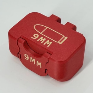 Puede incluir: Una caja roja rectangular con tapa abisagrada. La caja presenta el texto "9MM" y un gráfico de una bala en color claro. La caja está diseñada para guardar munición.