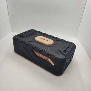 May include: A black rectangular container with a tan-coloured centre panel that reads "9MM." The container has a textured surface and a sliding mechanism on the side. The text "8777310" is printed on the side.