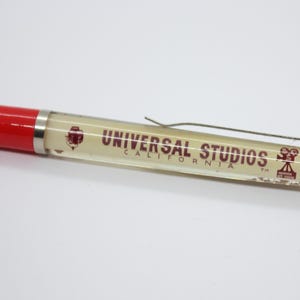 May include: A red and clear pen with the text "UNIVERSAL STUDIOS CALIFORNIA" printed on the barrel. The pen has a silver clip and a red tip. The pen is a souvenir.