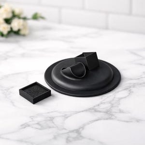 May include: A set of black, minimalist home decor items. The set includes a round tray with a geometric accent, a small square coaster with a pattern, and a folded object. The items are arranged on a white marble surface.