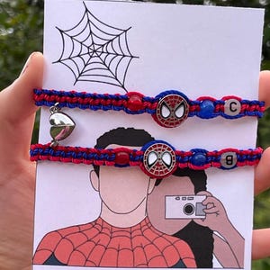 Könnte beinhalten: Zwei passende rot-blaue gewebte Armbänder mit Spider-Man-Emblemen und Buchstabenperlen. Ein Armband hat einen silbernen Herzanhänger. Die Armbänder werden auf einer Karte mit einer Spider-Man-Illustration und einem Spinnennetz präsentiert.