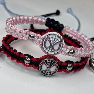 May include: Close-up of two friendship bracelets. One bracelet is pink and white, the other is red and black. Both bracelets have a silver charm with a Spider-Man design. The bracelets are made of woven cord and have adjustable closures.