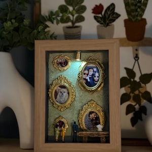 Może przedstawiać: Pudełko cieniowe z jasną drewnianą ramą, zawierające kilka małych, ozdobnych, złotych ramek z zdjęciami. Tło to teksturowana zielona tkanina. Wewnątrz pudełka znajdują się również miniaturowe figurki i małe światło. Pudełko stoi na drewnianej półce.