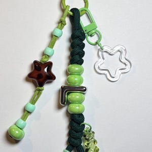 May include: A keychain featuring a dark green braided cord and a light green suede cord. The design includes light green beads, a brown star-shaped bead, a silver letter L charm, a clear star-shaped charm, and a small translucent green bear charm.
