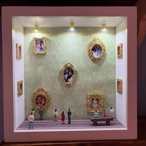 Puede incluir: Una vitrina blanca con retratos en miniatura enmarcados y pequeñas figuras. Los retratos tienen marcos dorados y están montados en una pared con dibujos verdes. La exhibición tiene un suelo de madera y está iluminada por pequeños focos.