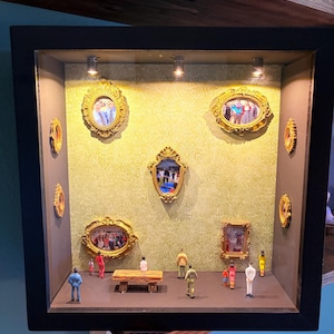 Puede incluir: Un diorama en miniatura en un marco negro, con un fondo de pantalla verde y varias imágenes pequeñas con marco dorado. Pequeñas figuras están dispuestas en una plataforma, con un pequeño banco. El diorama está iluminado por pequeños focos.