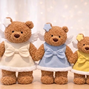 May include: Four brown teddy bears with angel wings and star-shaped headbands. Each bear wears a dress in a different pastel color: cream, blue, yellow, and pink. The dresses have a bow at the waist and white trim.