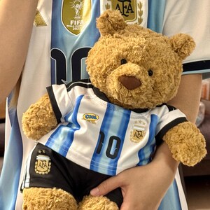 May include: A light brown plush teddy bear wearing a white and light blue striped soccer jersey with the number 10, the AFA logo, and a small oval patch reading COOL, paired with black soccer shorts featuring the AFA logo and two gold stars.