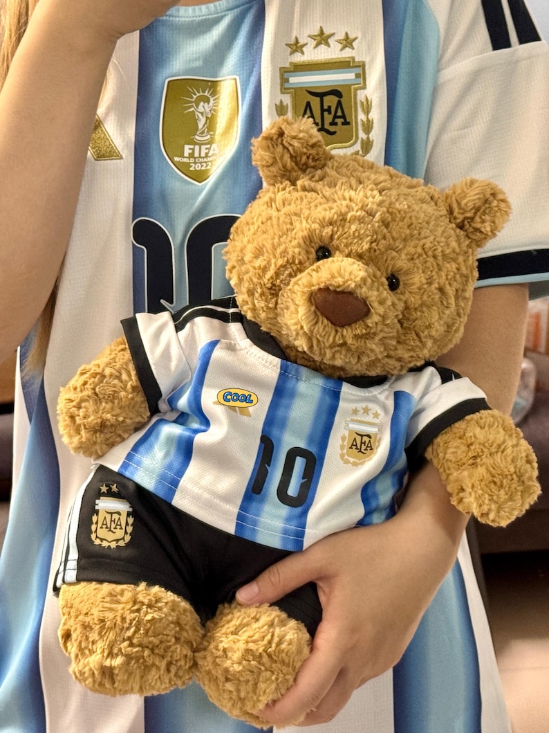 May include: A light brown plush teddy bear wearing a white and light blue striped soccer jersey with the number 10, the AFA logo, and a small oval patch reading COOL, paired with black soccer shorts featuring the AFA logo and two gold stars.