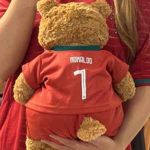 May include: A tan plush teddy bear wearing a red soccer jersey and matching shorts. The jersey features the name RONALDO and the number 7 printed in white on the back. The bear has a dark green collar and is made of soft, textured fur material.