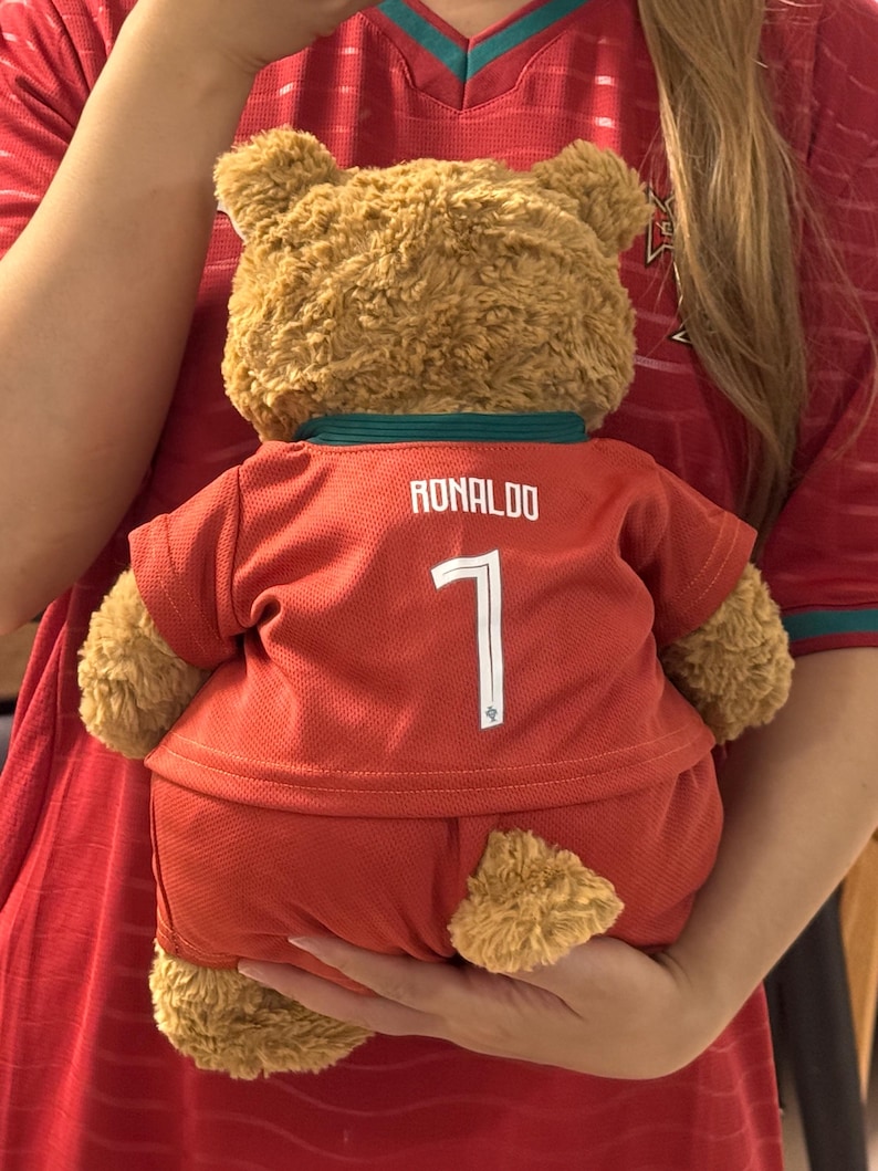 May include: A tan plush teddy bear wearing a red soccer jersey and matching shorts. The jersey features the name RONALDO and the number 7 printed in white on the back. The bear has a dark green collar and is made of soft, textured fur material.