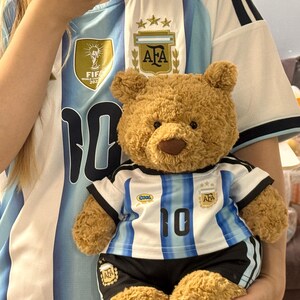 May include: A light brown plush teddy bear wearing a miniature soccer jersey and shorts set. The jersey features white and light blue vertical stripes, the number 10, an AFA crest with three stars, and a small blue and yellow 'cool' patch on the chest.