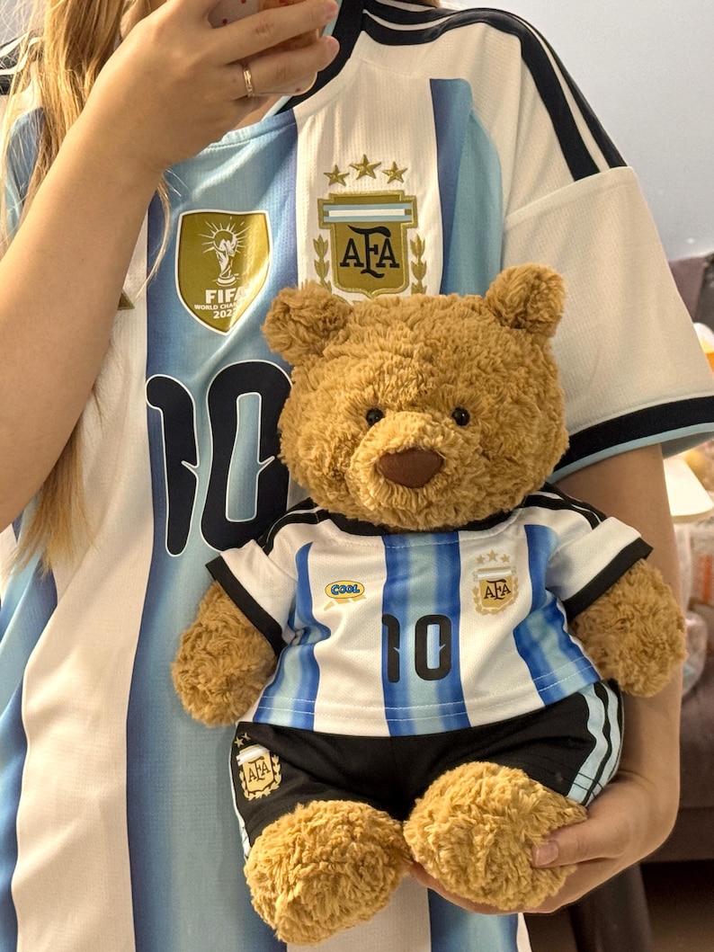 May include: A light brown plush teddy bear wearing a miniature soccer jersey and shorts set. The jersey features white and light blue vertical stripes, the number 10, an AFA crest with three stars, and a small blue and yellow 'cool' patch on the chest.