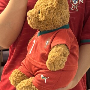 May include: A brown plush teddy bear wearing a red soccer jersey and matching red shorts. The jersey features a green collar, a white number 7, and a crest on the chest. The shorts display a white logo with the word COOL printed in small letters.