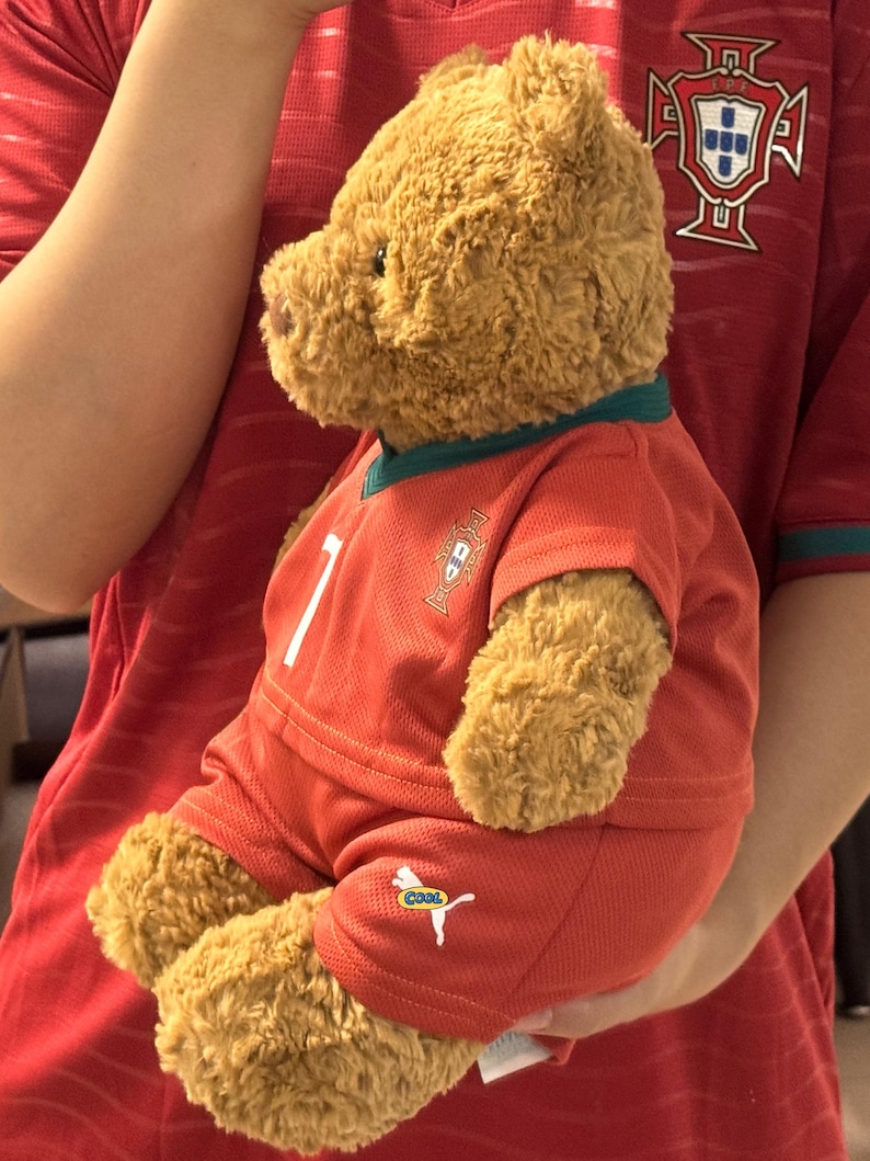 May include: A brown plush teddy bear wearing a red soccer jersey and matching red shorts. The jersey features a green collar, a white number 7, and a crest on the chest. The shorts display a white logo with the word COOL printed in small letters.