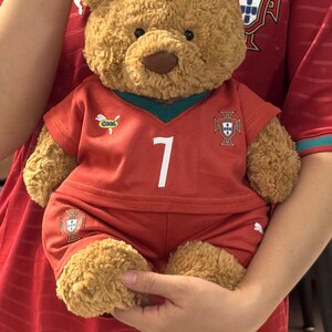 May include: A brown plush teddy bear wearing a red Portugal soccer jersey and matching shorts. The jersey features the number 7 in white, a small Puma logo, a 'COOL' patch, and the Portugal national team crest on the chest and shorts.
