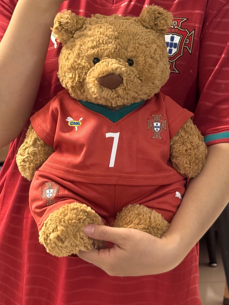 May include: A brown plush teddy bear wearing a red Portugal soccer jersey and matching shorts. The jersey features the number 7 in white, a small Puma logo, a 'COOL' patch, and the Portugal national team crest on the chest and shorts.