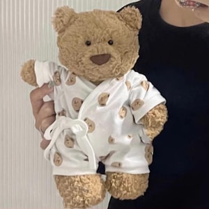May include: A brown teddy bear wearing a white robe patterned with small brown bear faces. The robe is tied with a white sash. The bear has a brown nose and stitched mouth. The toy is a soft, cuddly item.