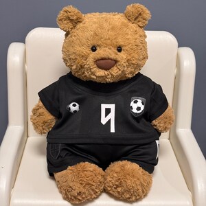 May include: A plush brown teddy bear wearing a black soccer jersey and matching black shorts. The jersey features a white number 9 on the front, a soccer ball graphic on the chest, and a small soccer ball emblem on the side of the shirt.