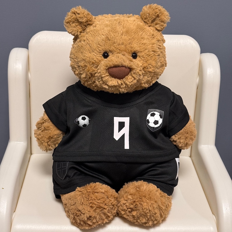 May include: A plush brown teddy bear wearing a black soccer jersey and matching black shorts. The jersey features a white number 9 on the front, a soccer ball graphic on the chest, and a small soccer ball emblem on the side of the shirt.