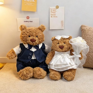 May include: Two brown teddy bears, one in a navy blue suit and bow tie, the other in a white wedding dress and veil, holding a bouquet. The bears have tan snouts and paws. Text on the wall.