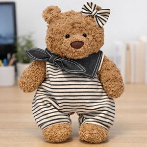 May include: A plush teddy bear with brown fur, wearing a white and black striped romper and a dark denim collar. The bear has a black and white striped bow on its head, a dark brown nose, and black eyes, standing in an upright position.