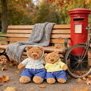 May include: Two brown teddy bears wearing blue jeans and hoodies, one gray and one yellow, sit on a walkway in front of a park bench. A gray blanket rests on the bench. A red mailbox and a bicycle are in the background.
