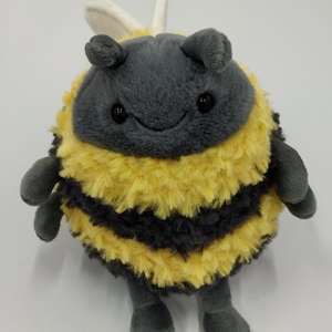 Puede incluir: Un juguete de peluche con forma de abeja con un cuerpo redondo, con rayas amarillas y negras. La abeja tiene alas, antenas y pies pequeños grises. El juguete tiene una cara sonriente con ojos negros. Este peluche es adecuado para niños.