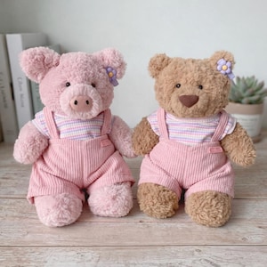 Puede incluir: Dos peluches: un cerdo rosa y un oso marrón. Ambos visten monos de pana rosa y camisas a rayas. Cada uno tiene un pequeño accesorio floral. El cerdo tiene un hocico rosa y el oso una nariz marrón.