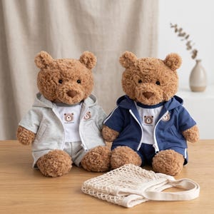 Peut inclure: Deux ours en peluche marron portant des tenues assorties composées d'un t-shirt blanc avec un petit motif d'ours, d'un short et d'un sweat à capuche zippé. L'un porte un sweat gris clair, l'autre un sweat bleu marine. Un sac en filet blanc est posé devant.